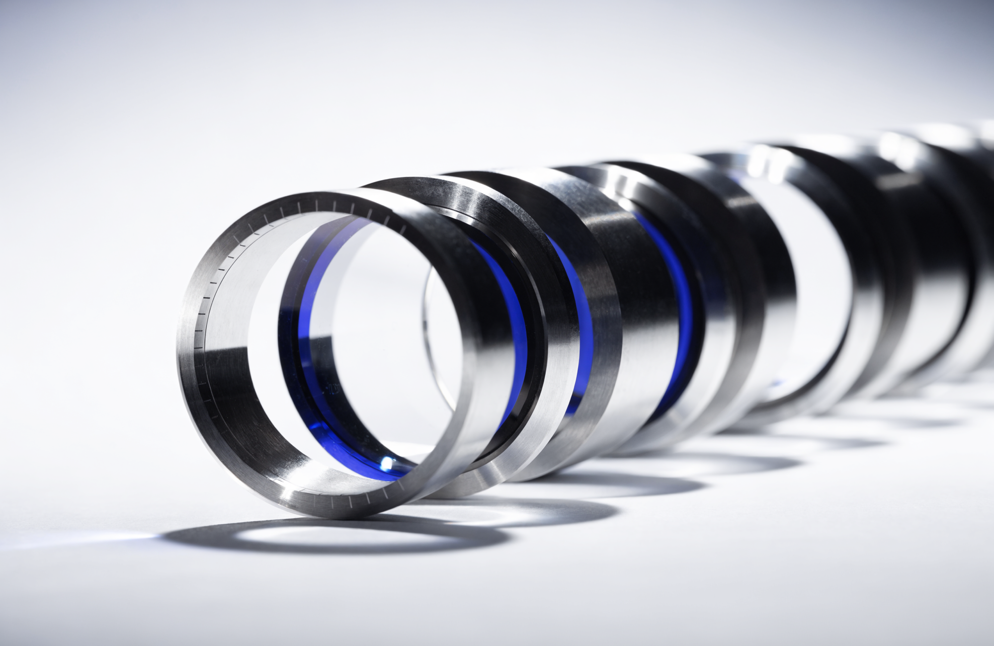 A row of precision machined lens rings receding into the distance, the nearest tack-sharp with a cobalt-blue core of light, the rest softening out of focus.